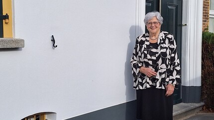 Vrouw met grijs haar staat voor een donkergrijze voordeur van een wit huis. Ze draagt een zwart-witte blouse met bladvormig patroon en een zwarte rok.