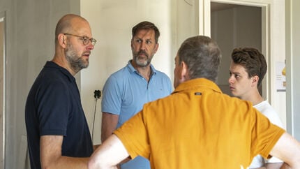 Vier personen staan in gesprek in een lichte ruimte met houten elementen en onafgewerkte muren, tijdens een bouwgerelateerde bijeenkomst.